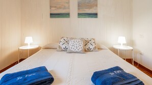Quarto de hotel com uma cama grande, duas mesas de cabeceira, duas luminárias fixadas na parede e duas pinturas de uma praia.