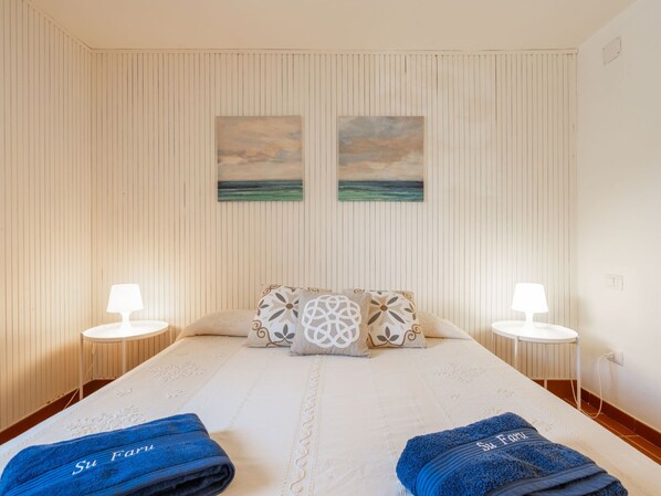 Una habitación de hotel con una cama grande, dos mesitas de noche, dos lámparas de pared y dos cuadros con una playa.