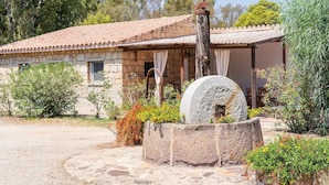 Une maison en pierre avec un toit de tuiles, un puits en pierre et une végétation luxuriante tout autour.