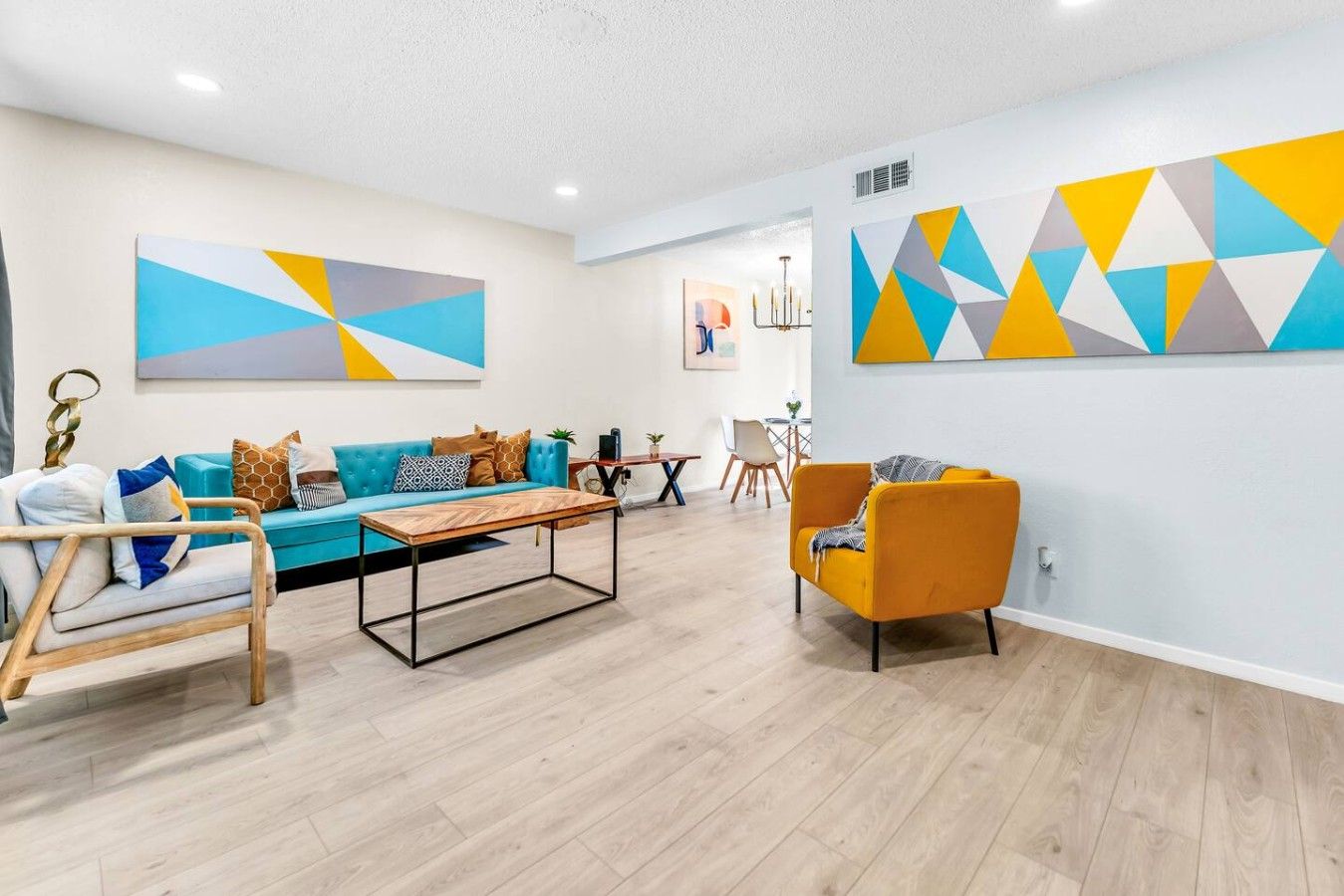 A modern living room with a sofa, coffee table, and armchairs, featuring geometric wall art and a chandelier.