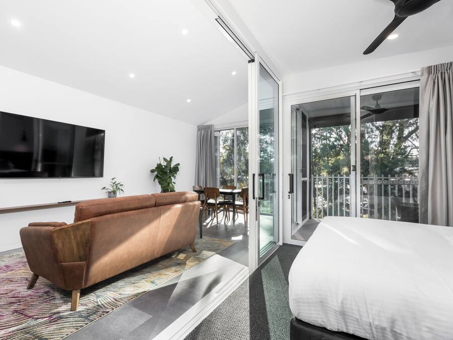A modern hotel room with a large bed, a sofa, a TV, a balcony with a view, and a dining area.