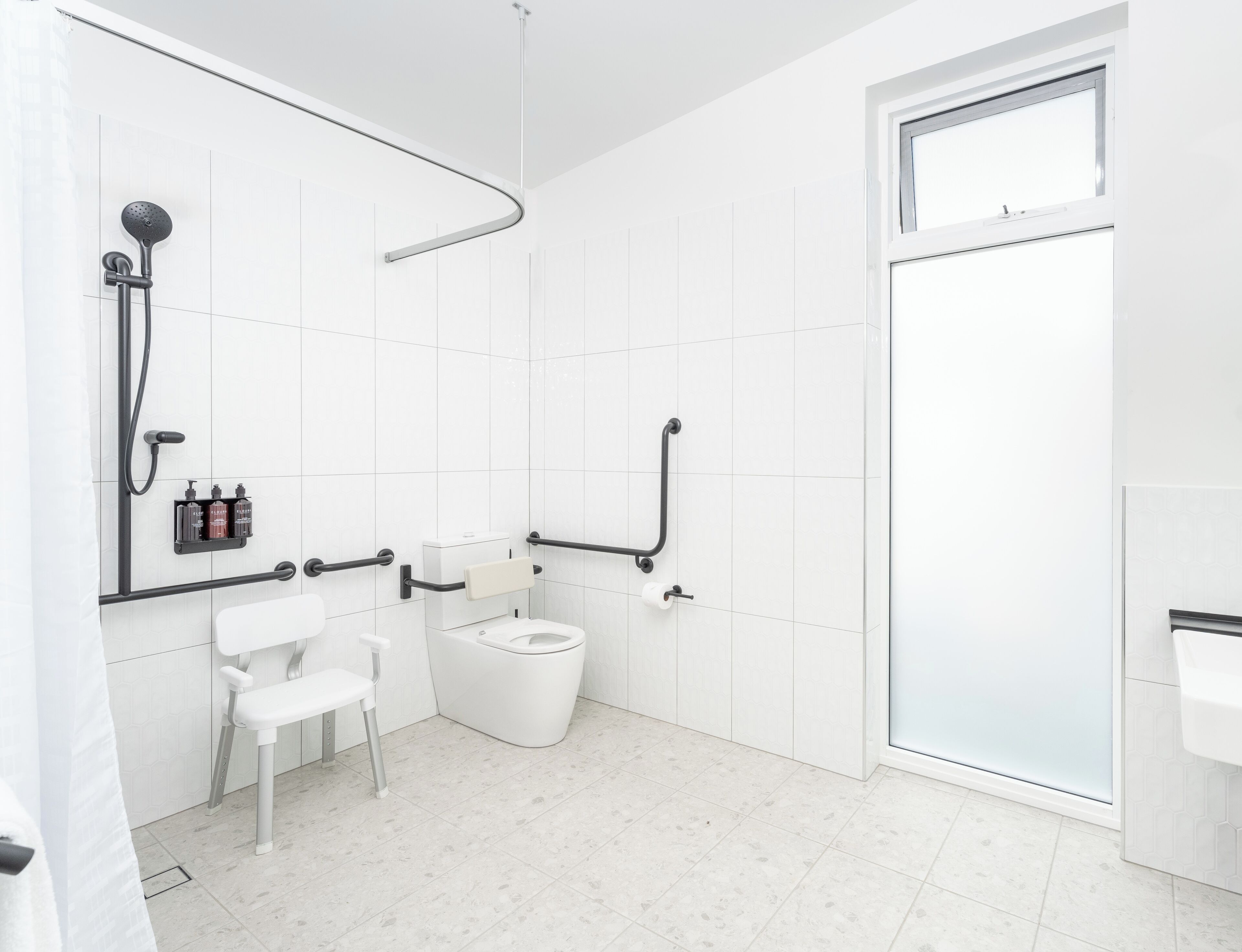 Een moderne badkamer met witte tegels, een douche met een gebogen douchekop, een toilet met een handgreep en een witte stoel.