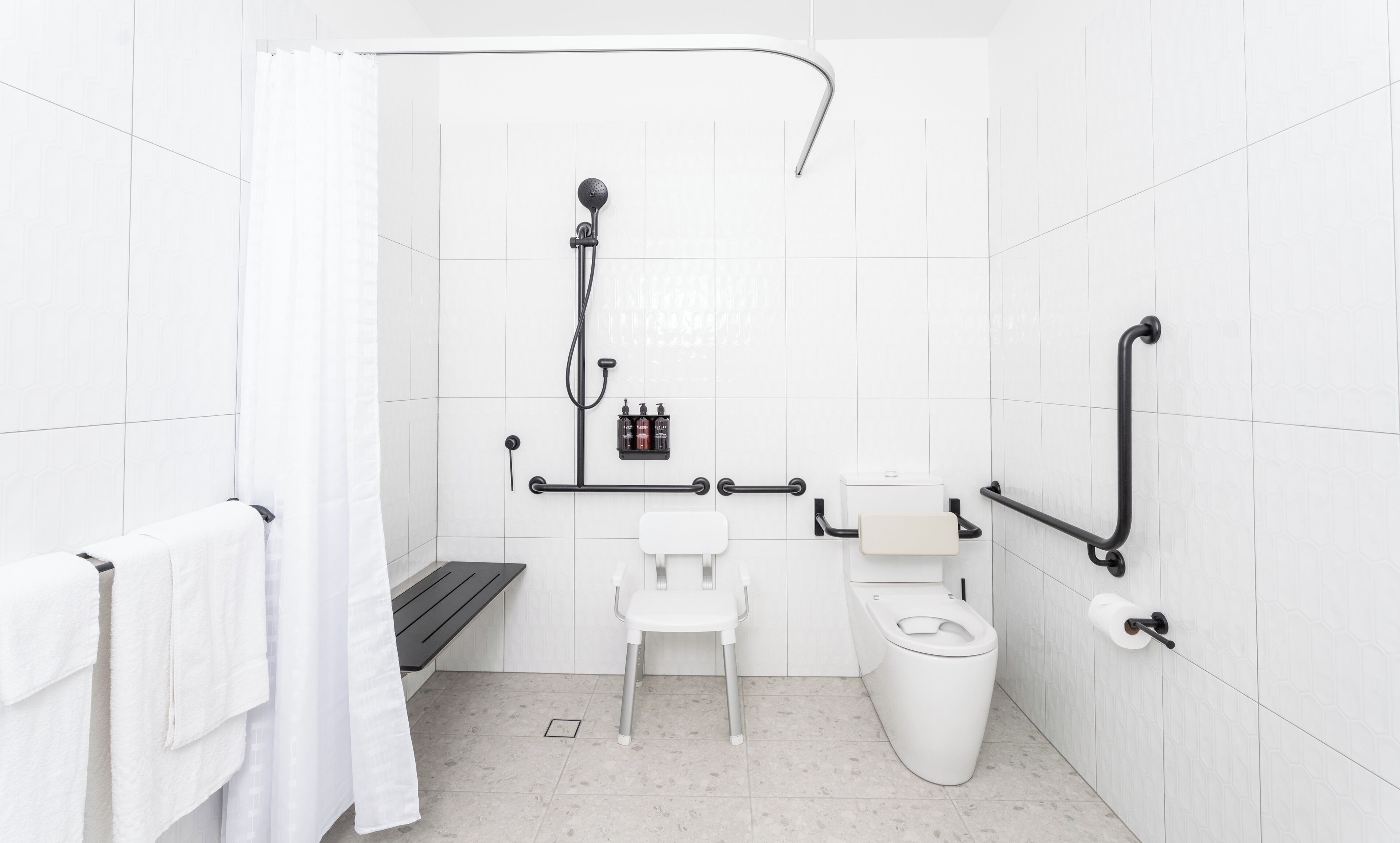 Un baño moderno con azulejos blancos, una ducha con cabezal de ducha manual, un inodoro, un banco y barras de apoyo.