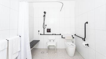 Un baño moderno con azulejos blancos, una ducha con cabezal de ducha manual, un inodoro, un banco y barras de apoyo.