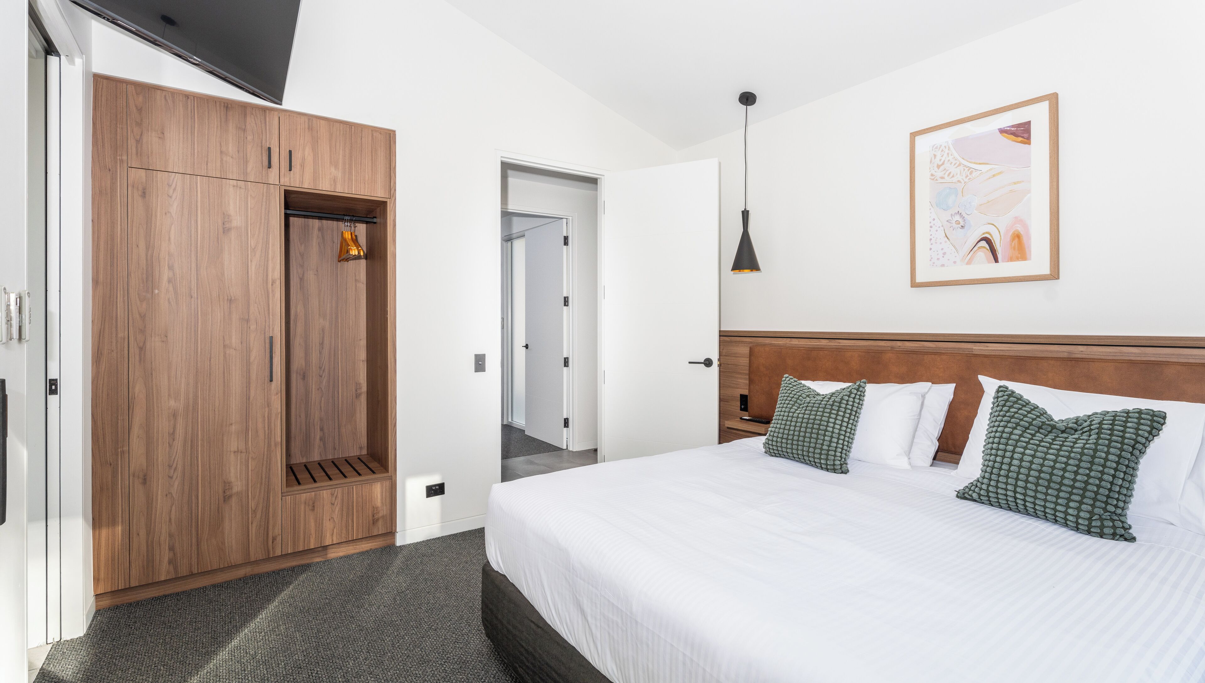 Une chambre d’hôtel avec une armoire en bois, un lit agrémenté d’oreillers à motifs verts, une œuvre d’art encadrée au mur et un luminaire suspendu.