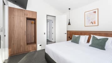 A hotel room with a wooden wardrobe, a bed with green patterned pillows, a framed artwork on the wall, and a pendant light.