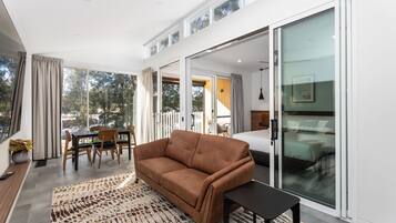 A modern living room with a sofa, a small table, and a dining area with chairs. There is a large window providing a view of the outdoors.