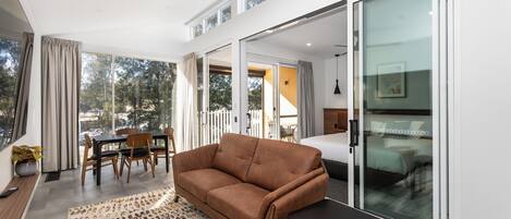 A modern living room with a sofa, a small table, and a dining area with chairs. There is a large window providing a view of the outdoors.