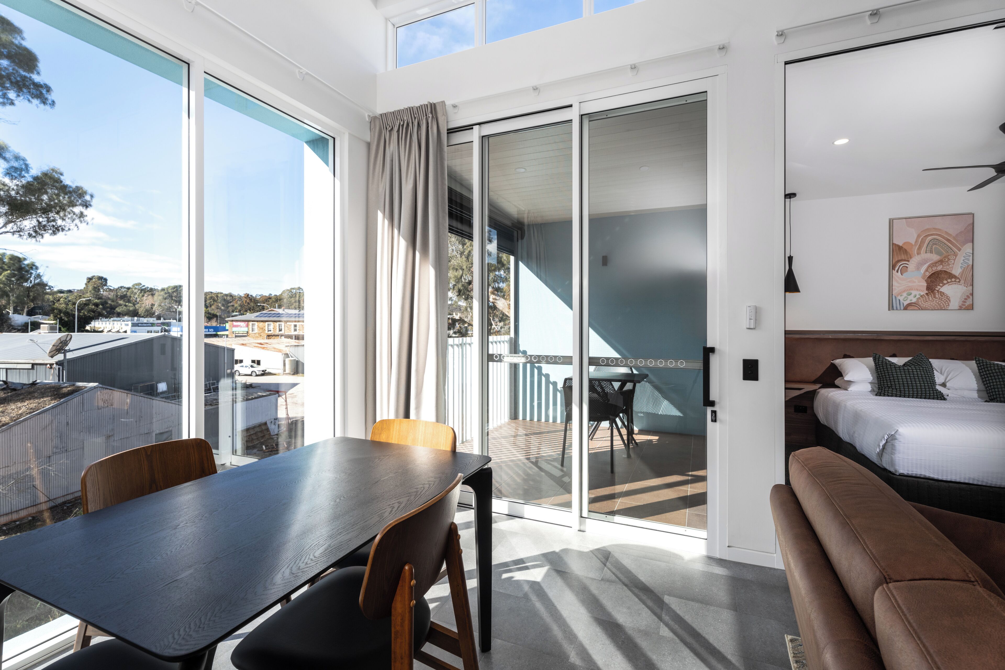 A modern hotel room with a large sliding glass door leading to an outdoor balcony, a wooden dining table, a sofa, and a bed with a headboard.