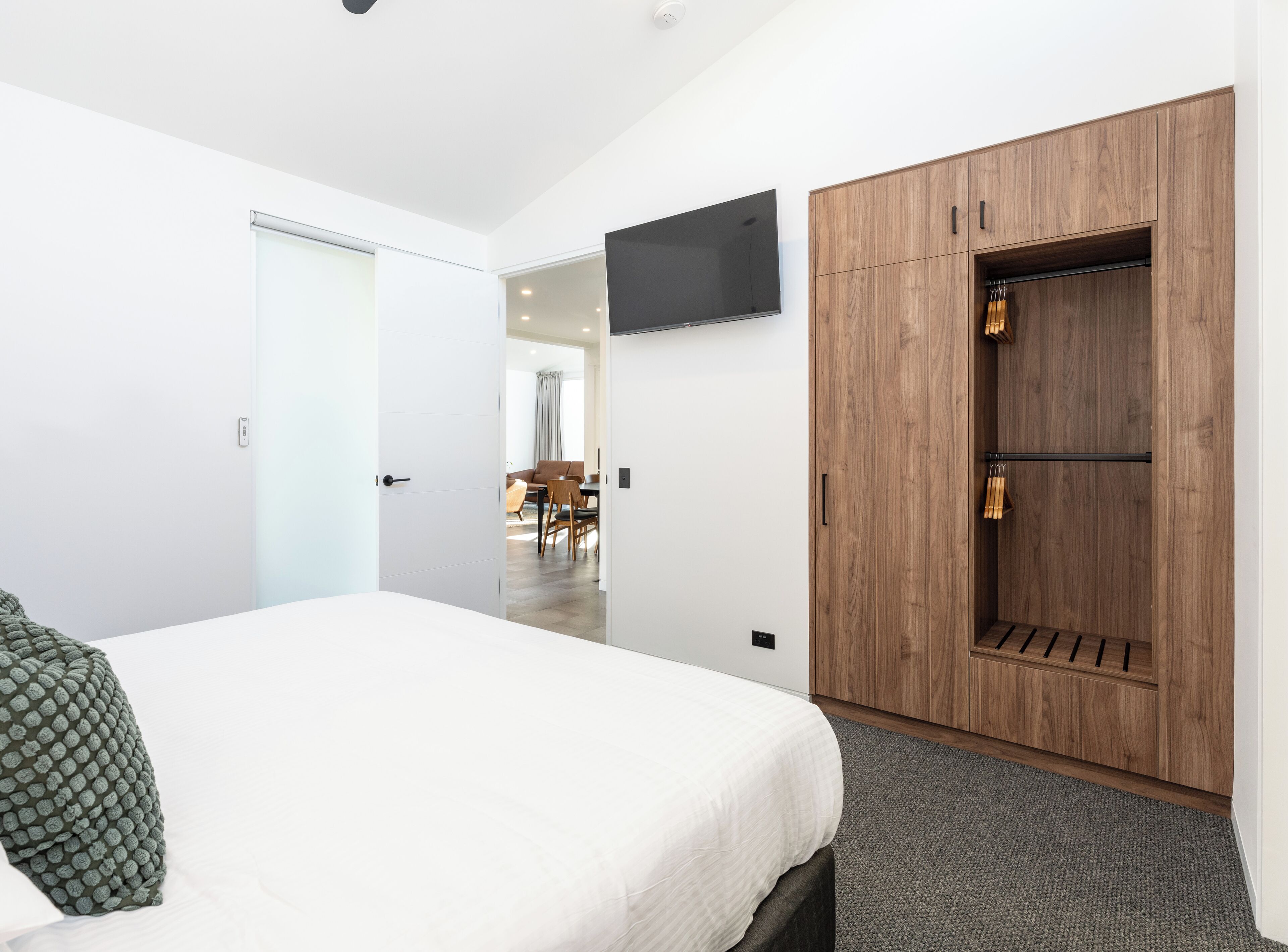 A modern hotel room with a large bed, a wooden wardrobe, a flat-screen TV, and a dining area visible through an open door.