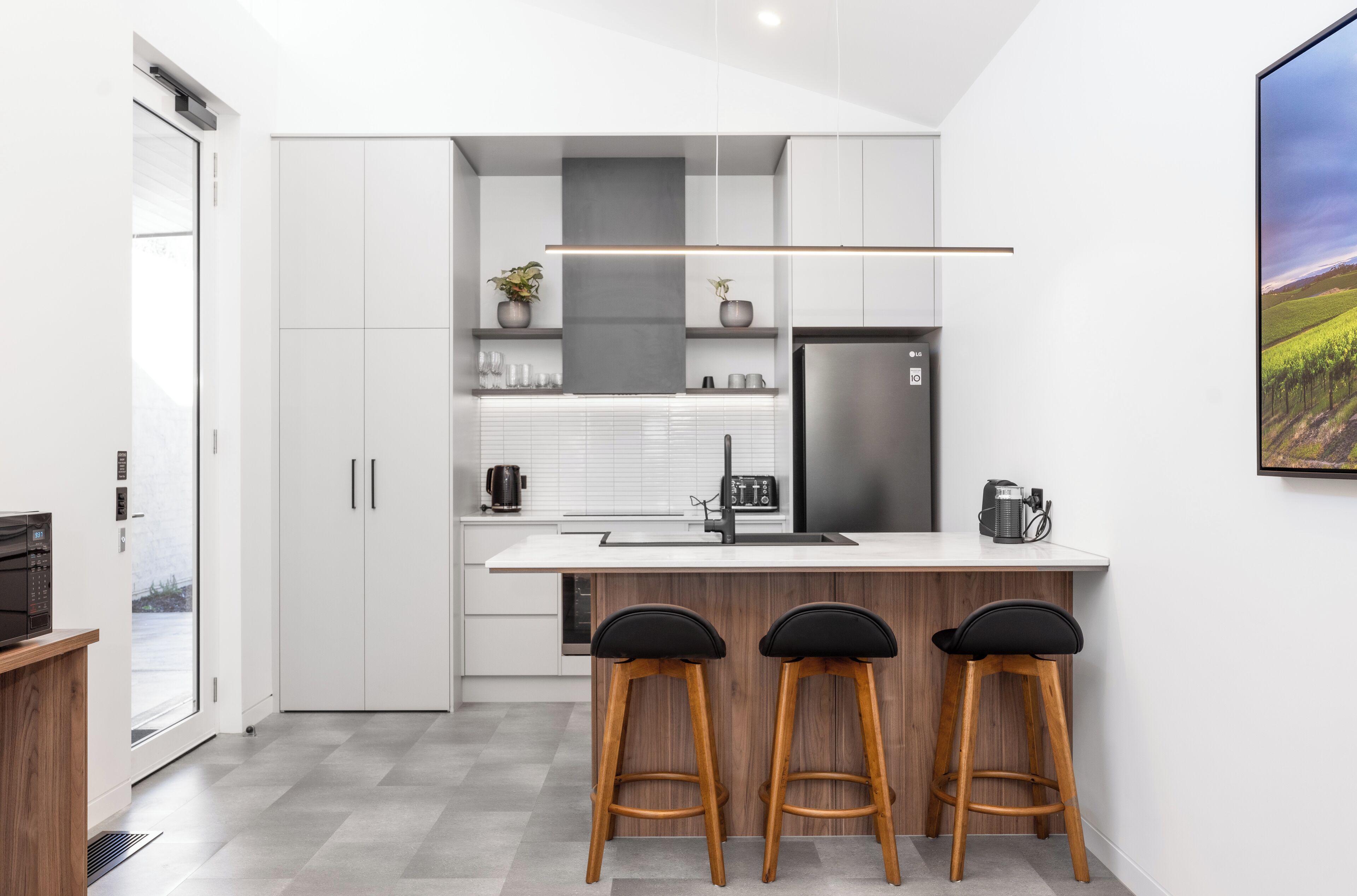 A modern kitchen with white cabinetry, a stainless steel refrigerator, a white countertop, and three black bar stools.