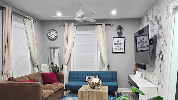 A modern living room with a blue sofa, a brown sofa, a wooden coffee table, a black dining table with chairs, a ceiling fan, and a TV mounted on the wall. - Cozy 2BR Home in Delmar Loop (University City)