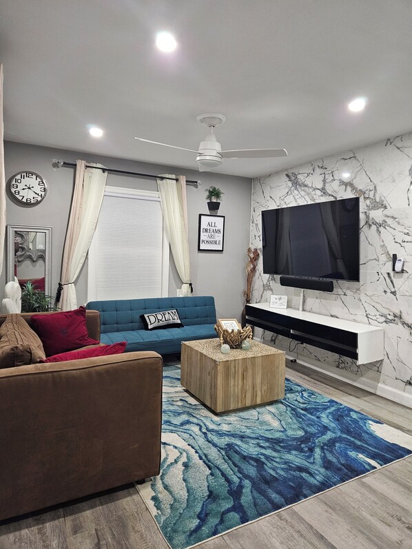 A modern living room with a blue sofa, a brown sofa, a wooden coffee table, a blue patterned rug, a flat-screen TV, a ceiling fan, a wall clock, and a motivational quote on the wall.