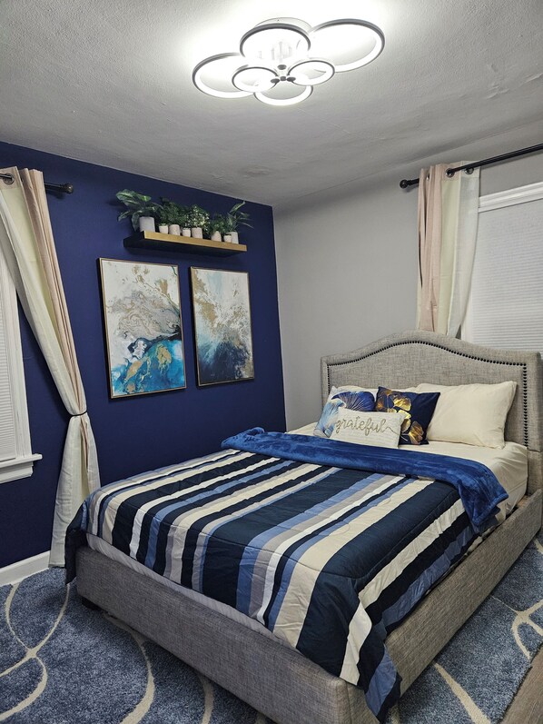A bedroom with a bed, a ceiling light fixture, a wall-mounted shelf with plants, and two framed artworks. - Cozy 2BR Home in Delmar Loop (University City)