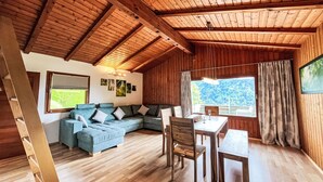 Ein gemütliches Blockhaus-Interieur mit Holzbalkendecke, einer Couch, einem Esstisch und Blick ins Freie.