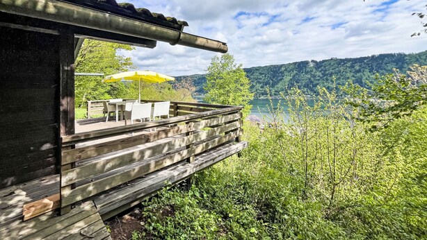 Eine Holzterrasse mit Sitzgelegenheit und einem gelben Sonnenschirm mit Blick auf einen See und bewaldete Hügel.