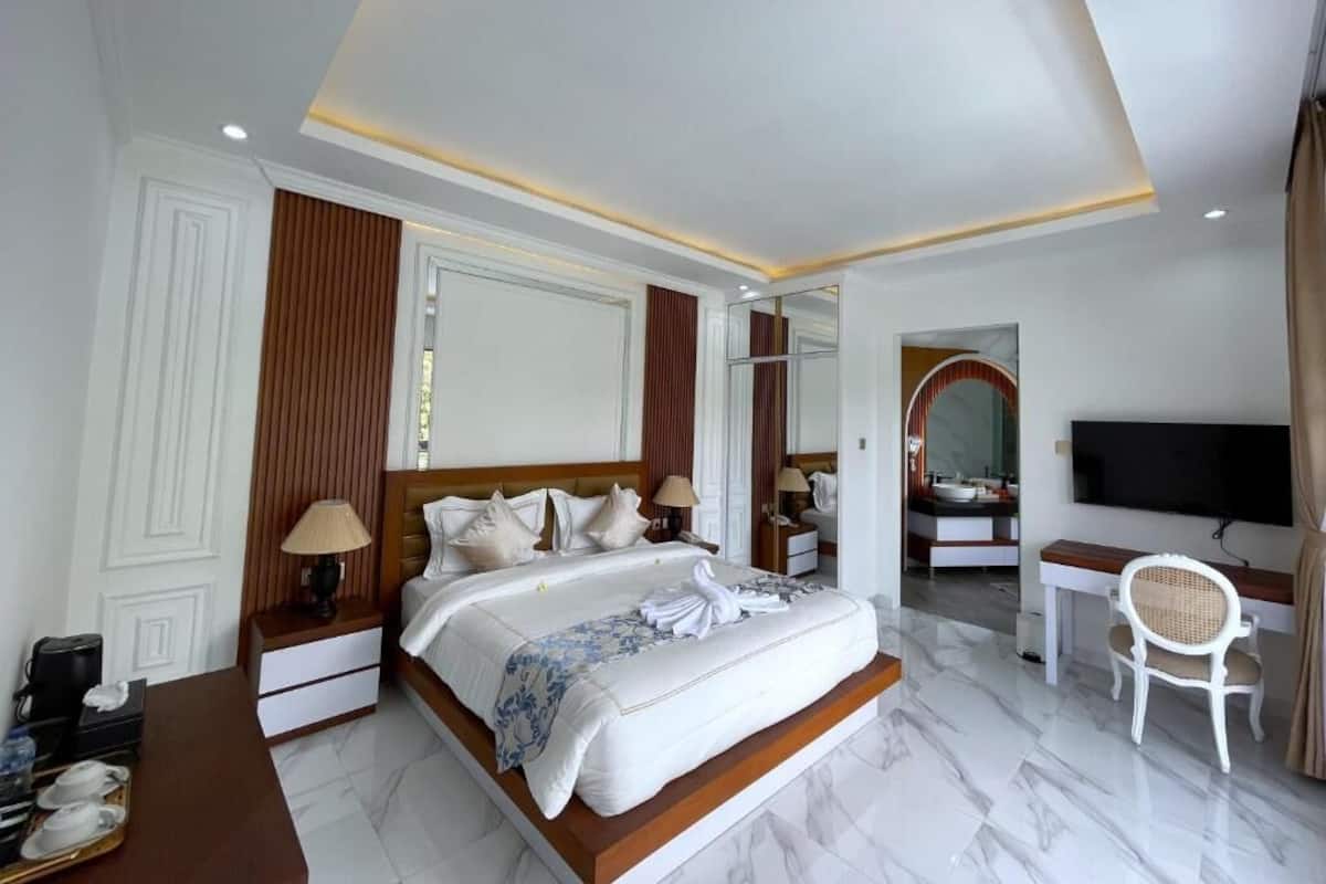 A hotel room with a large bed, a desk, a chair, a TV, and a bathroom visible through an archway.