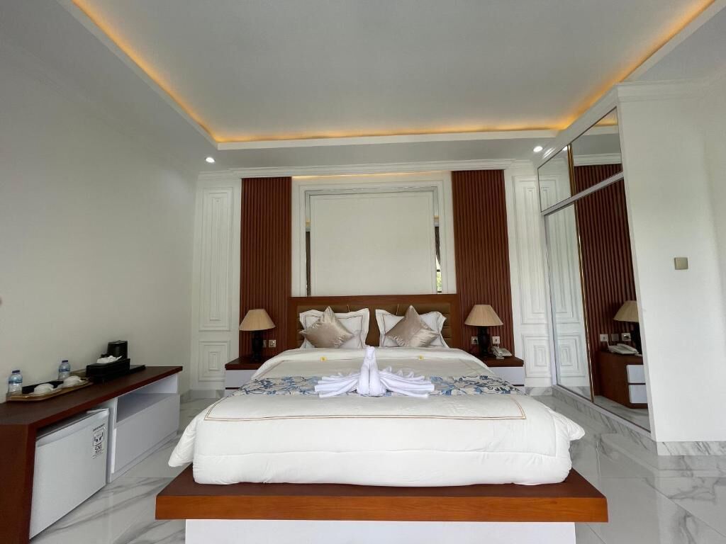 A hotel room with a large bed, a wooden headboard, two bedside tables with lamps, a mirror, and a small desk with a chair and a water bottle.