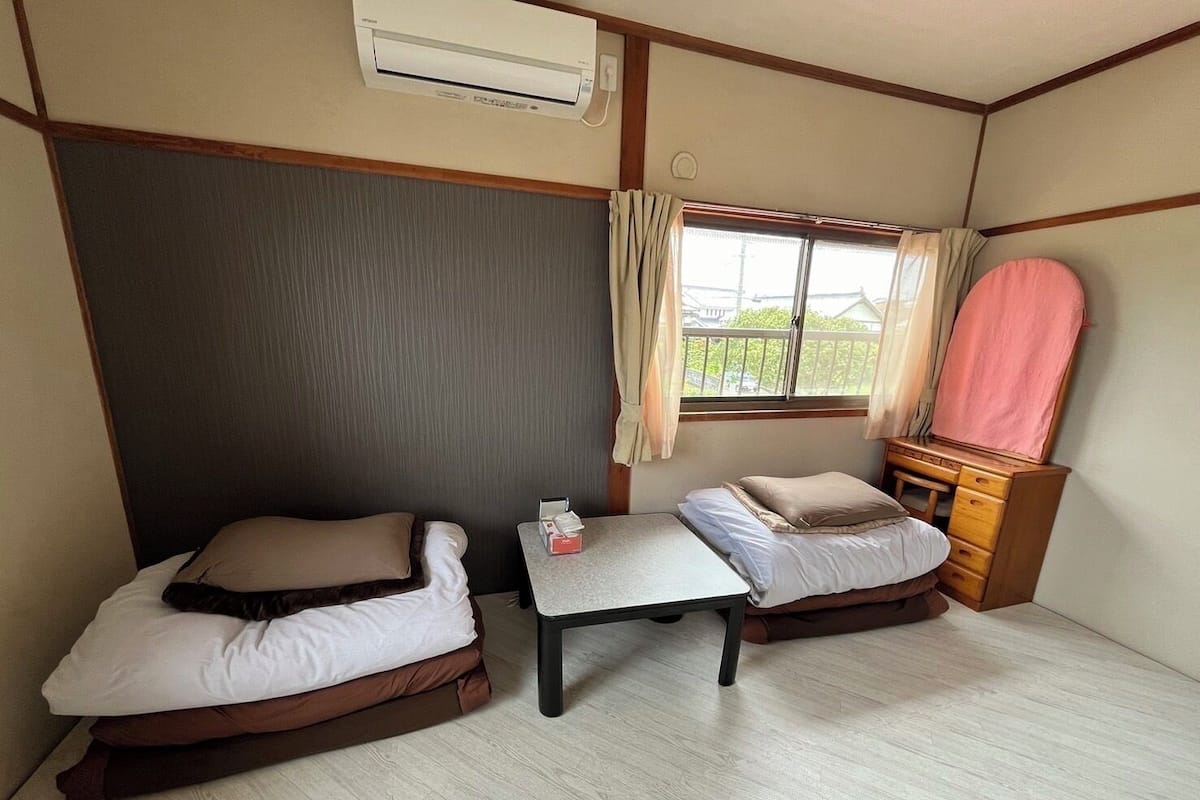 A compact room with two beds, a small table, a dresser, and an air conditioning unit.
