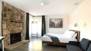 Habitación de hotel con chimenea de piedra, cama con cabecera de madera, sofá gris y un cuadro en la pared.