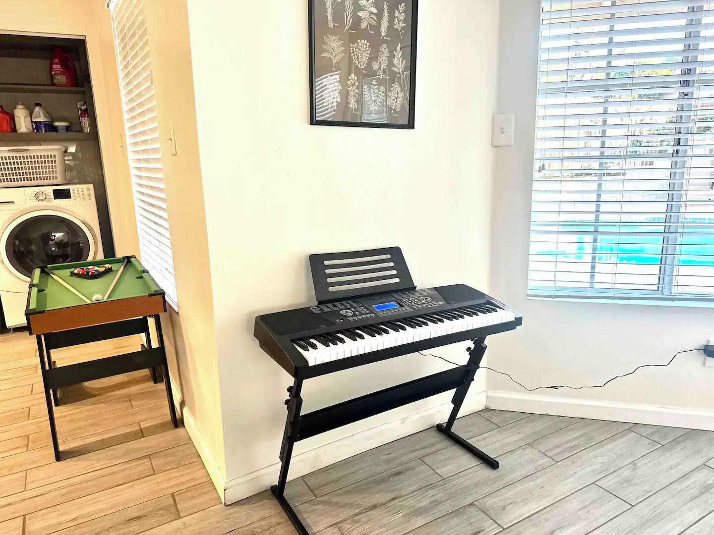 Una habitación con mesa de billar, lavadora, teclado y ventana con persianas.
