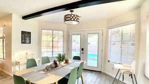 Un comedor con mesa de madera, sillas verdes, lámpara colgante y ventanas amplias con persianas.