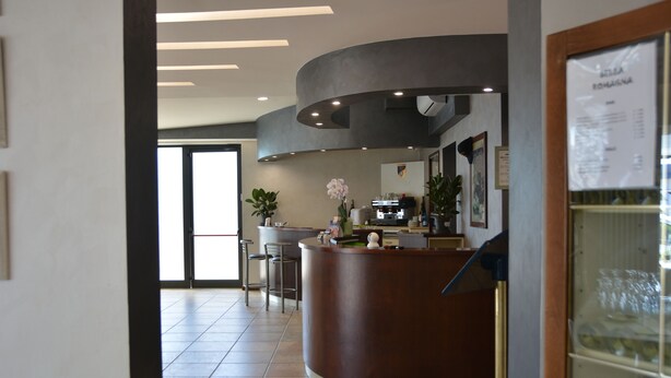 A modern reception area with a curved counter, a seating area with chairs, and a view of the kitchen in the background. - Hotel  Bella  Romagna  (Ravenna)