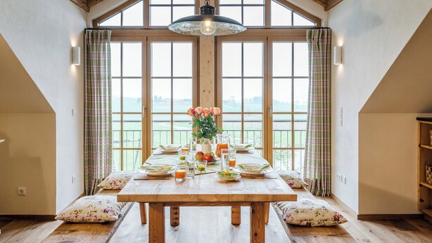 Une table à manger dressée pour le petit-déjeuner, avec vue sur l’extérieur, des poutres en bois au plafond et une grande fenêtre avec des rideaux.