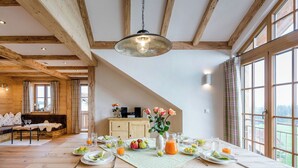 Un coin repas avec une table en bois mise pour le petit-déjeuner, un canapé et une vue sur l’extérieur grâce à de grandes fenêtres.