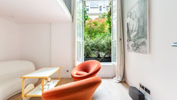 Een moderne woonkamer met een witte bank, een houten bijzettafeltje en een unieke oranje fauteuil. Een groot raam biedt uitzicht op een groene tuin.