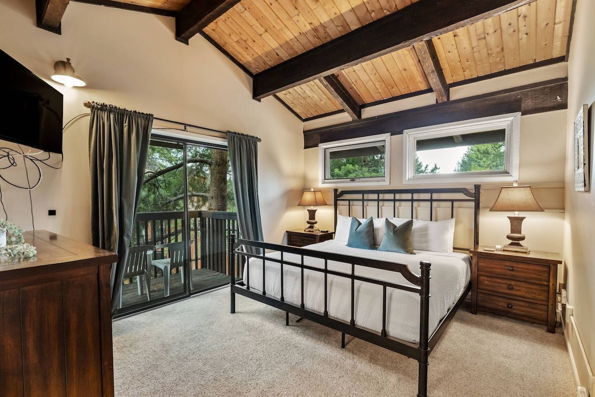 A bedroom with a bed, two bedside lamps, a wooden headboard, a window with curtains, a desk with a lamp, and a view of greenery outside.