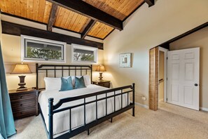 Un dormitorio con techo de madera, una cama con ropa blanca, dos lámparas de noche y una puerta a otra habitación.