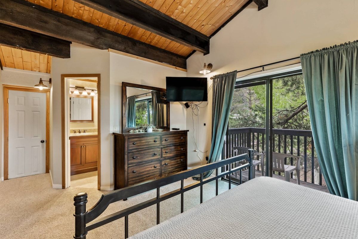 Une chambre avec un grand lit, une commode, une télévision et un balcon donnant sur les arbres.