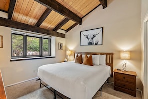 A bedroom with a large bed, wooden ceiling beams, a window with a view, and a nightstand with a lamp.