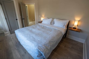 Un dormitorio con una cama grande, dos mesitas de noche con lámparas, suelo de madera y una puerta que da a otra habitación.