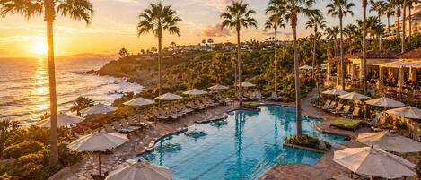 A resort pool with sun loungers and umbrellas, surrounded by palm trees and overlooking the ocean.