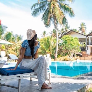 A person sitting on a lounge chair by a pool with palm trees and a house in the background. - Samudra Beach Resort (Rekawa)