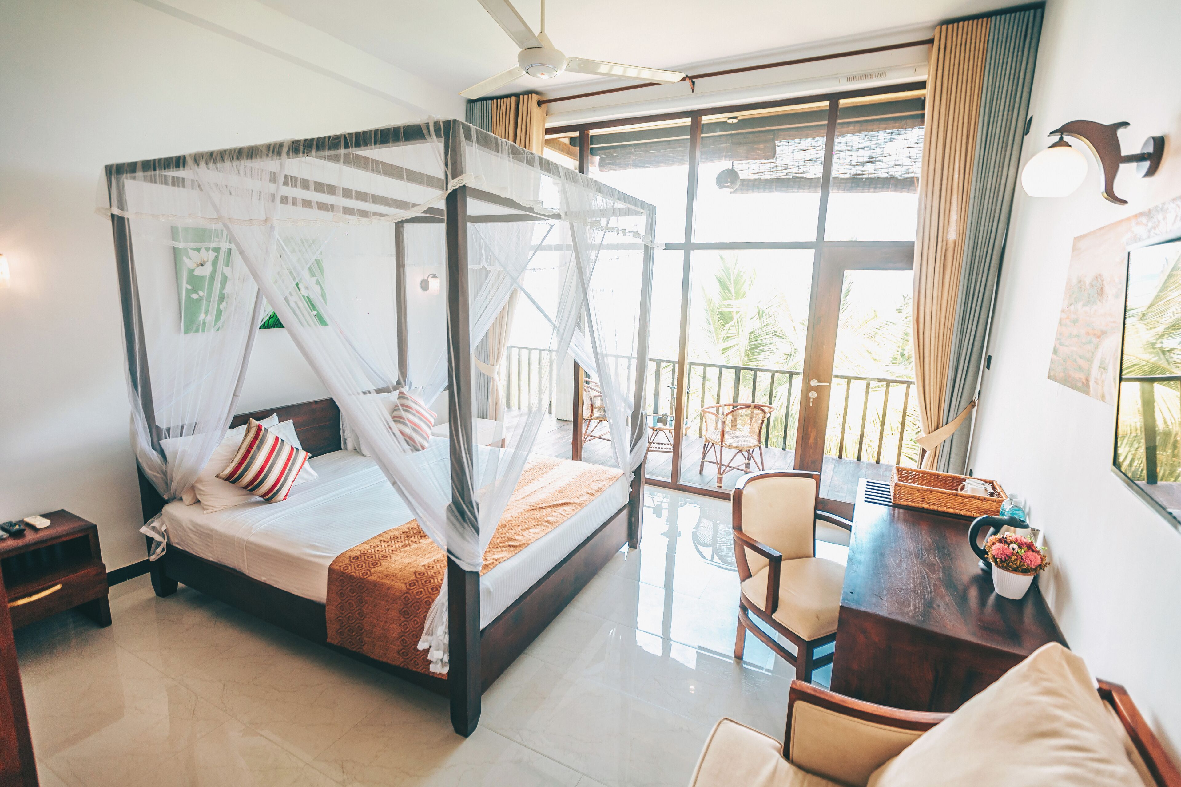A four-poster bed with a canopy, a dining table with chairs, and a balcony with a view of palm trees.