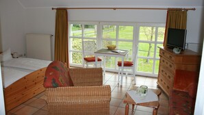 Een slaapkamer met een bed, een rieten stoel, een klein tafeltje met een schaal en glazen, en een dressoir met een televisie.