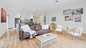 Ein modernes Wohnzimmer mit einer braunen Ledersofa, weißen Sesseln und einem Holztisch. Im Hintergrund ist eine Küche mit weißen Schränken und Edelstahlgeräten zu sehen.
