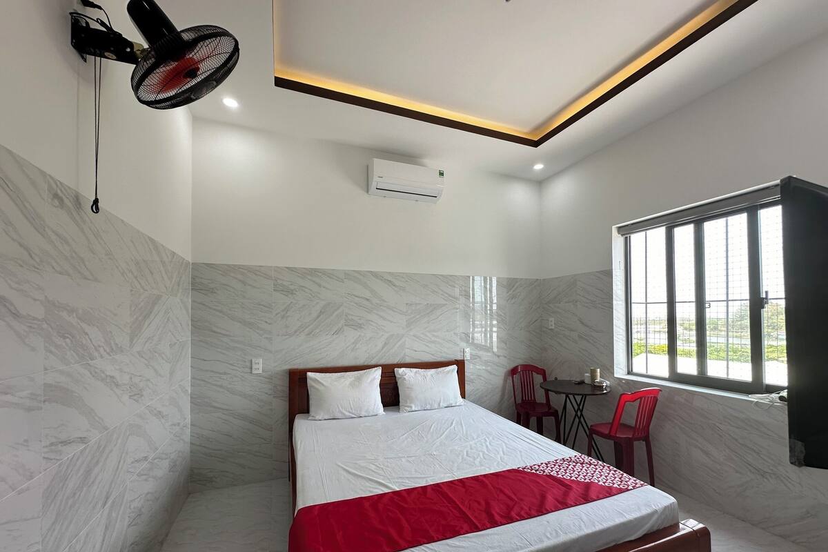 A hotel room with a bed, a red chair, a table, and a window with bars.