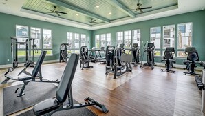 Ein großzügiger Fitnessraum mit diversen Trainingsgeräten, großen Fenstern und einer hohen Decke mit Ventilatoren.