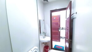 A small, compact bathroom with a toilet, sink, and a mirror. A red bucket is placed near the door, and a welcome mat is visible at the entrance. - Do Gia Hotel (Ho Chi Minh City)