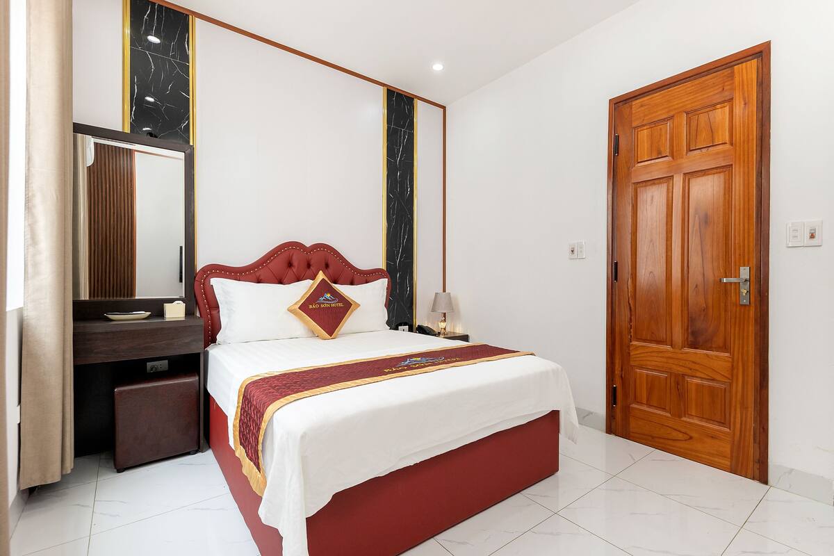 A hotel room with a bed, a bedside table, a mirror, and a wooden door.