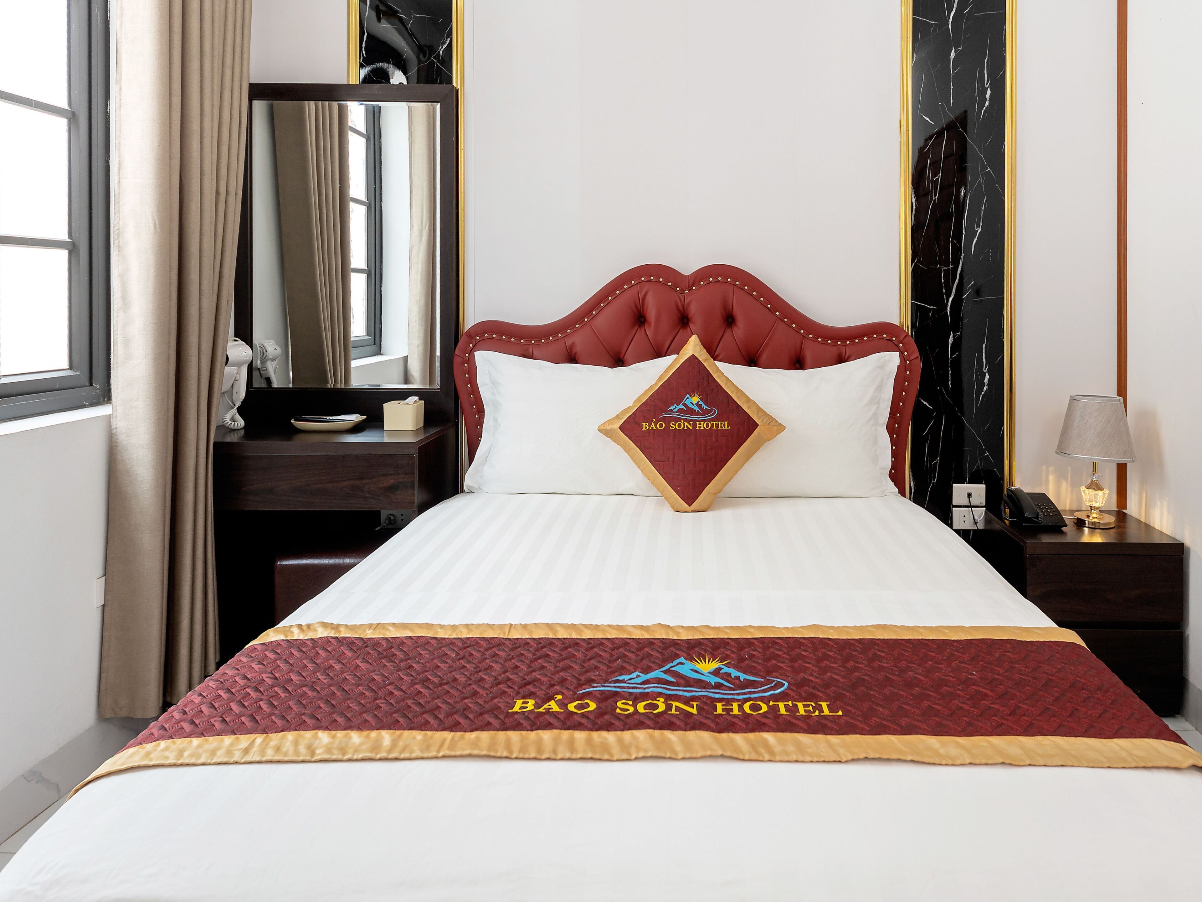 Quarto de hotel com cama, mesa de cabeceira, abajur, espelho e janela com cortinas.