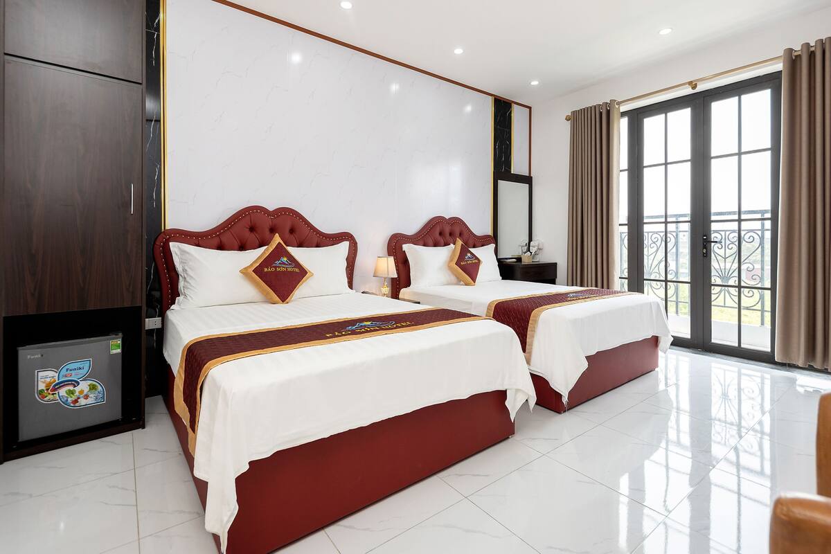 A hotel room with two beds, a minibar, a large window with curtains, and a view of the outside.