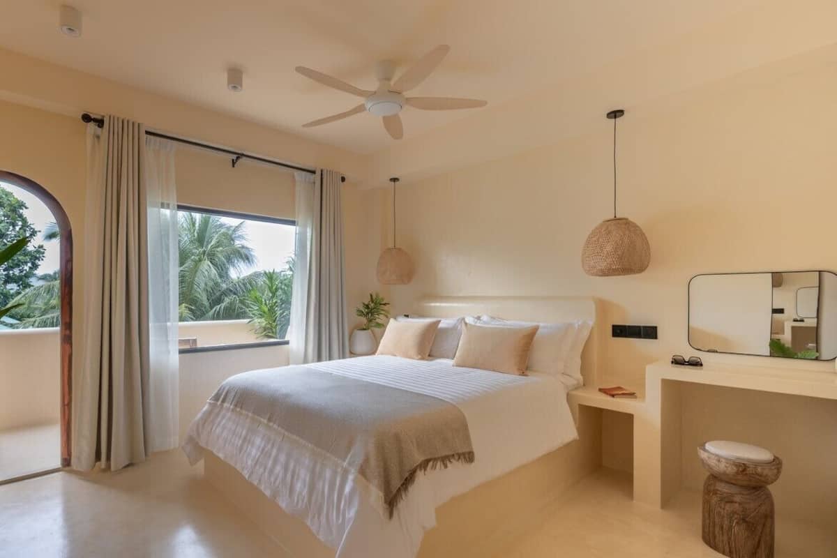 Une chambre avec un lit, un ventilateur de plafond, une table de chevet avec un miroir et une vue sur les palmiers par la fenêtre.
