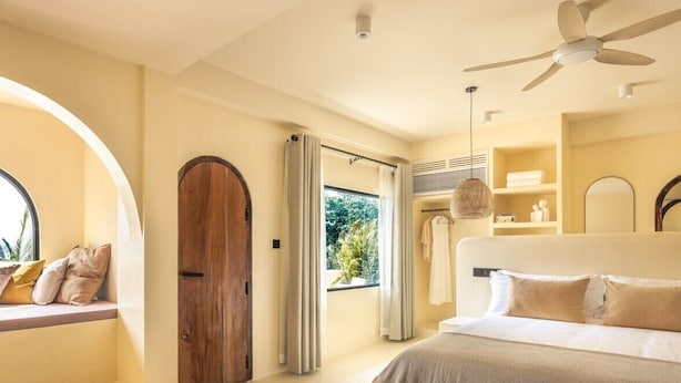 Ein Schlafzimmer mit einem großen Bett, einem Deckenventilator, einem Fenster mit Vorhängen und Blick ins Grüne.