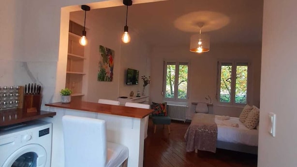A compact living space with a kitchen area, a washing machine, a dining table with chairs, and a bed in the background. - Amazonia Rouen Centre by Myfrenchpat (Rouen)