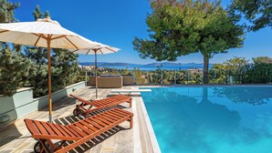 Un espace aménagé au bord de la piscine, avec des chaises longues et un parasol, entouré d’arbres et offrant une vue sur la mer.